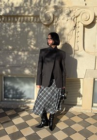 Femme avec un carré noir portant un blazer sombre, une jupe à carreaux, des bottes noires et des lunettes, debout contre un mur en pierre orné sous la lumière du soleil.