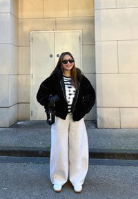 Chaqueta de pelaje sintético negro, camiseta a rayas con gráfico, pantalones de chándal grises de pierna ancha, gafas de sol negras y zapatillas blancas con suelas de goma.