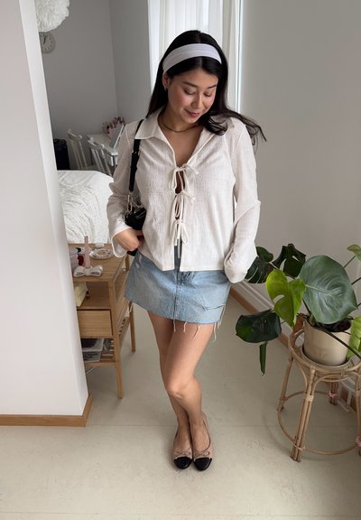 Blusa blanca de lino con detalle de cordones, combinada con una minifalda de mezclilla azul claro. Zapatos planos en negro y beige, y un bolso negro. Planta en maceta cerca.