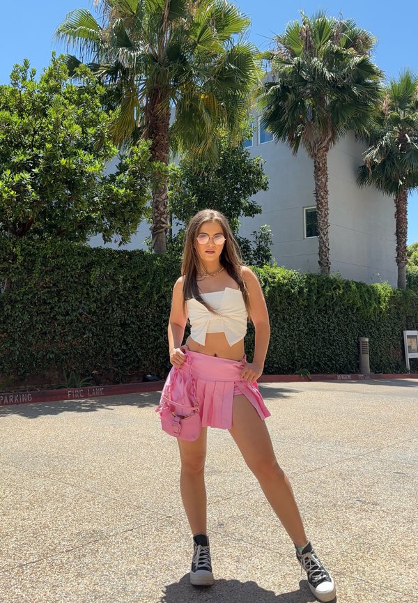 Valkoinen lyhyt toppi, vaaleanpunaiset laskostetut minihameet, mustat tennarit. Taustalla palmupuita ja vihreää kasvillisuutta. Kirkas auringonvalo.