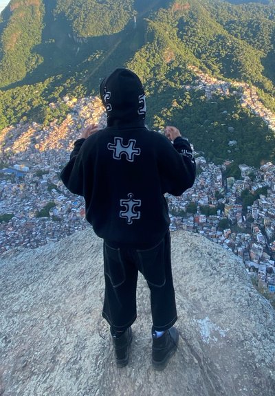 Sudadera negra con diseños de piezas de rompecabezas, pantalones cortos negros holgados y botas negras. La figura está de pie sobre una superficie rocosa con vista a una ciudad en la ladera.
