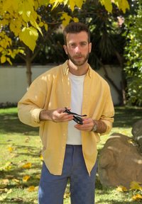 Άνδρας με κίτρινο πουκάμισο και μπλε παντελόνι, που κρατάει μαύρα γυαλιά ηλίου, στέκεται σε κήπο φωτισμένο από ήλιο με κίτρινα φθινοπωρινά φύλλα.