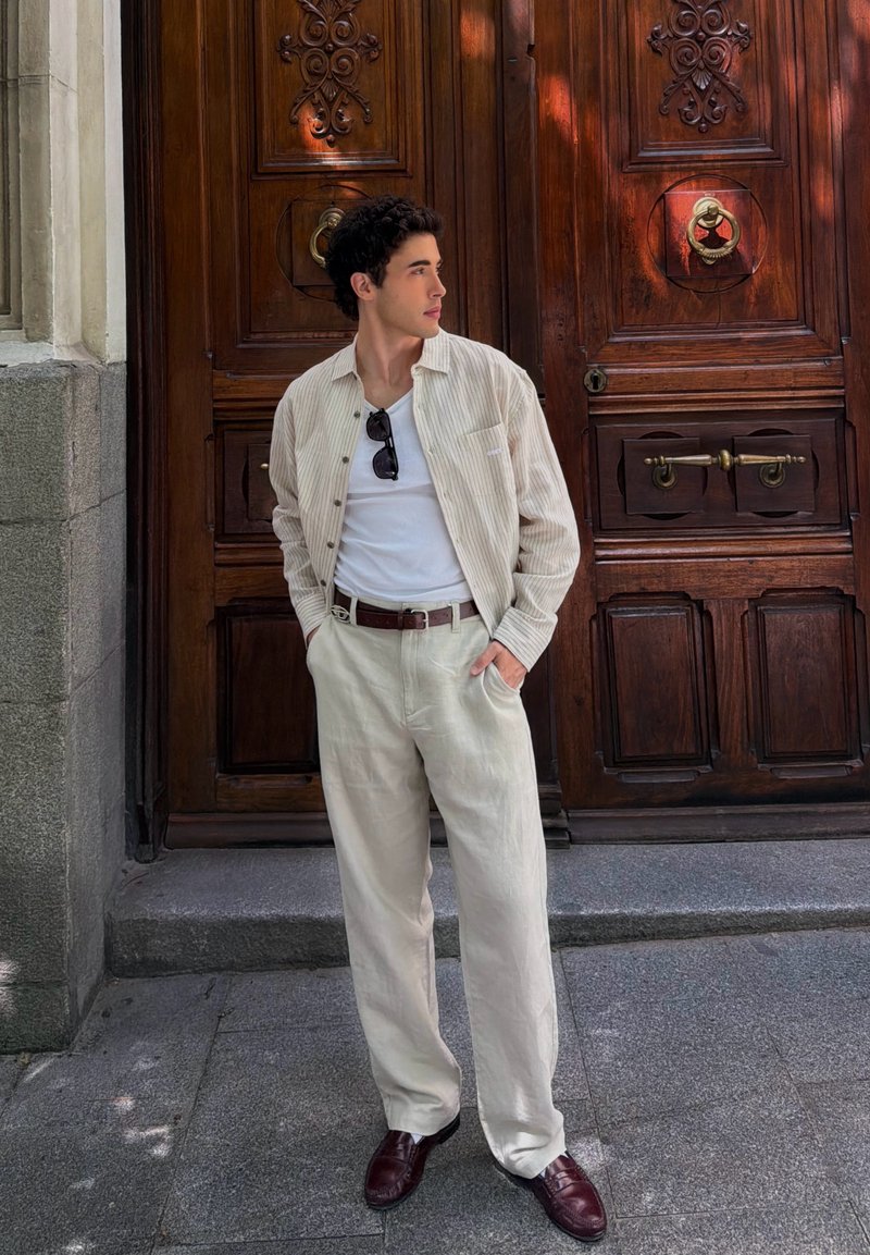 Jeune homme en pantalon beige clair et chemise rayée, les mains dans les poches, debout devant une grande porte en bois ornée, regardant sur le côté.