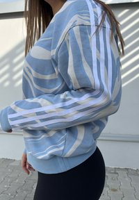 Light blue sweatshirt with white abstract patterns, featuring three white stripes along the sleeves and a relaxed fit. Textured cotton fabric.