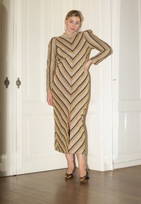 Robe dorée avec des rayures noires en chevrons, manches longues et fente haute sur la cuisse. Modèle portant des talons noirs et dorés, se tenant près d'un mur blanc.