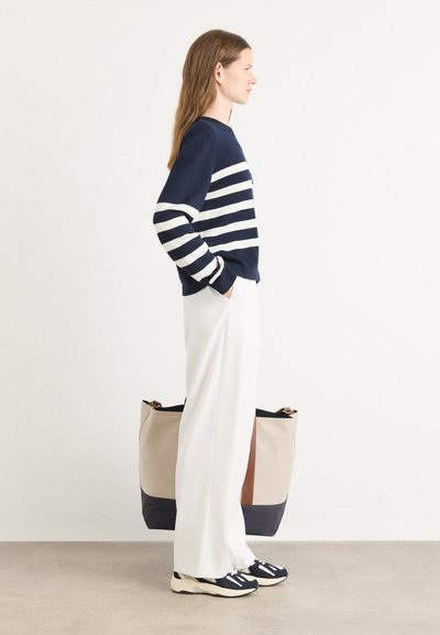 Suéter a rayas en azul marino y blanco, pantalones anchos blancos y zapatillas negras. Gran bolso de mano con acentos en beige, azul marino y marrón.
