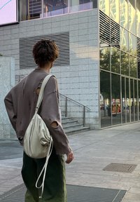 Rjavoverižna pulover z obrobnimi luknjami, v kombinaciji z zelenimi cargo hlačami. Bež torba z vrvico dopolnjuje oblačilo. Urbano okolje.