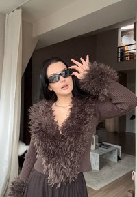 Pulover maro, cu dungi, având un guler și manșete din blană plușată, texturată; purtat cu ochelari de soare subțiri, de culoare închisă.