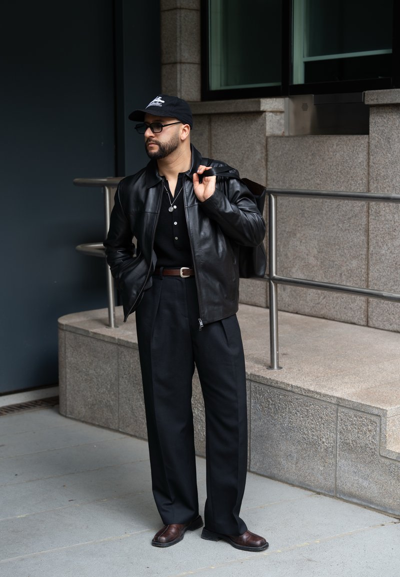 Homme portant une veste en cuir noire, une casquette noire, des lunettes de soleil, un pantalon noir, des chaussures marron, et portant un sac à dos sur une épaule dans un environnement urbain.