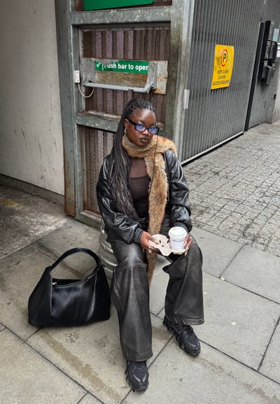Chaqueta oversized de cuero negro, bufanda de piel marrón, top negro, pantalones de cuero anchos, sosteniendo café; bolso negro texturizado y zapatillas en el pavimento.