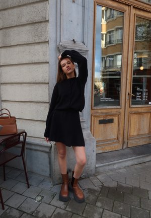 Vestido de punto negro con cierre frontal de botones, mangas largas y falda acampanada. La modelo lleva botas de plataforma de cuero marrón y tiene el cabello largo.