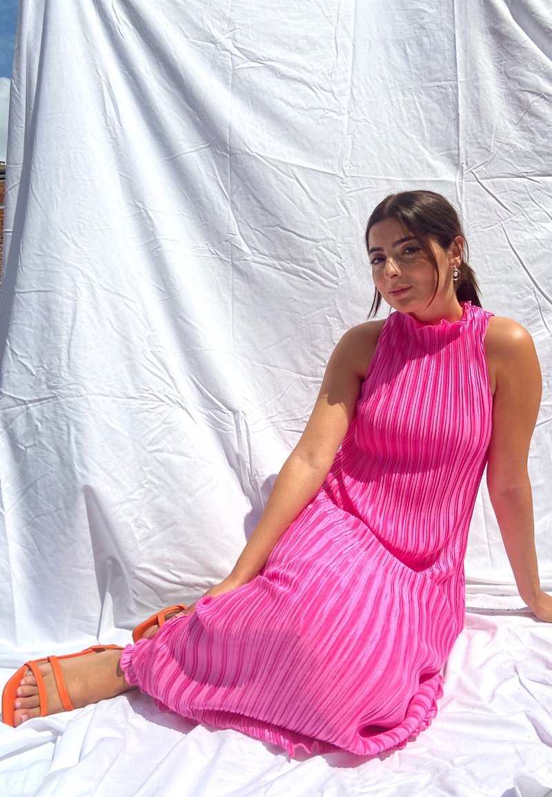 Fukszia redőzött ruha magas nyakkivágással, textúrált anyaggal és lágy sziluetttel, narancssárga pántos szandállal párosítva. Fehér háttér.