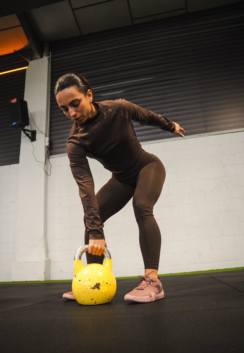 Kettlebell jaune avec une poignée métallique, posé sur un sol noir tandis qu'une personne vêtue de marron effectue un mouvement de levage.