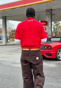 Červená tričko s krátkym rukávom s logom, biele tričko s dlhým rukávom pod ním, hnedé nohavice s náplasťou a oranžový opasok; v pozadí červené športové auto.