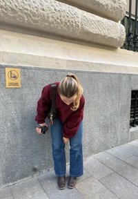 Suéter castanho, jeans azuis à boca de sino e botas castanhas; segurando uma câmara com uma bolsa. O fundo apresenta uma parede de pedra texturizada e um sinal de proibido fumar.