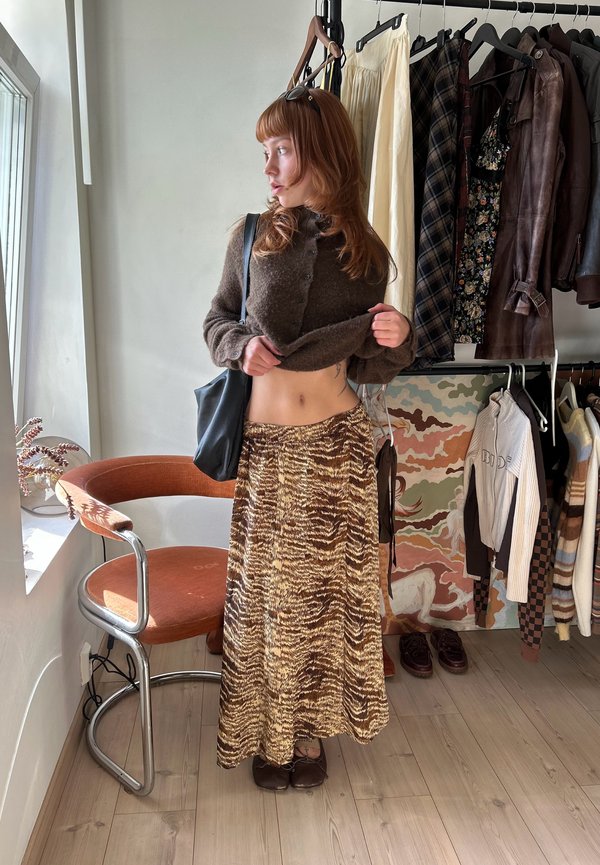 Γυναίκα με κόκκινα μαλλιά σηκώνει καφέ πουλόβερ, δείχνει τη μέση της, φορά μακρύ φούστα με μοτίβο τίγρης, στέκεται κοντά σε μια πορτοκαλί καρέκλα δίπλα σε μία κρεμάστρα ρούχων.
