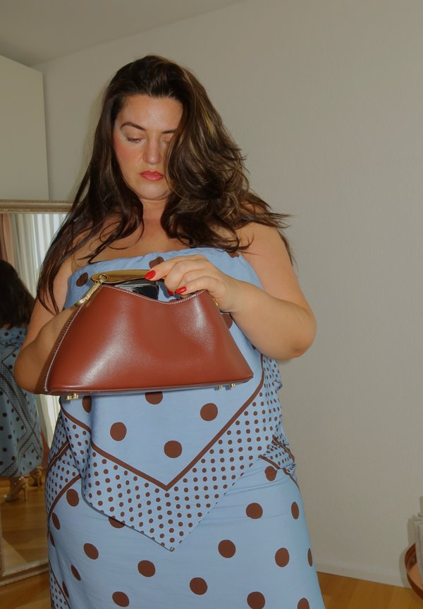 Femme aux longs cheveux bruns portant une robe bleue à pois tient et ouvre un sac à main en cuir marron à l'intérieur près d'un miroir.