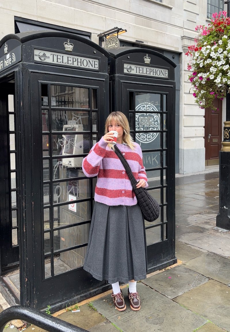 Cardigan lavorato a maglia a strisce rosa e rosse, gonna grigia plissettata, calze bianche, scarpe marroni. In piedi accanto a due cabine telefoniche nere.