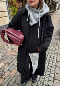 Manteau noir par-dessus un pull noir, écharpe grise, et un sac à main tissé bordeaux. Pantalon noir avec détail en dentelle. Fond de rue pavée.