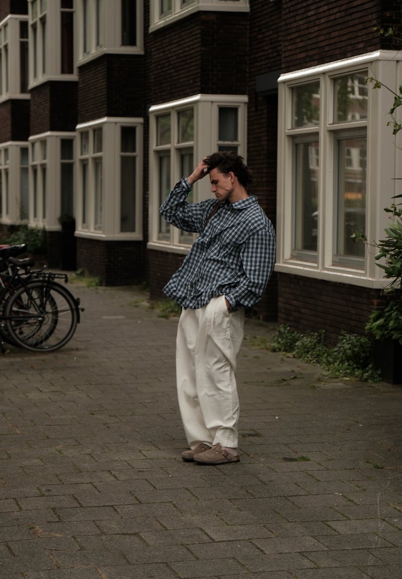 Homme en chemise à carreaux ample et pantalon blanc se tient sur le trottoir près des vélos et d'un bâtiment en briques aux fenêtres à encadrements blancs.