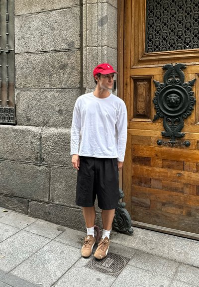 Un hombre está de pie contra una pared de piedra, vistiendo una camisa blanca de manga larga, pantalones cortos negros y zapatillas marrones con calcetines blancos. Detrás de él hay una puerta de madera.