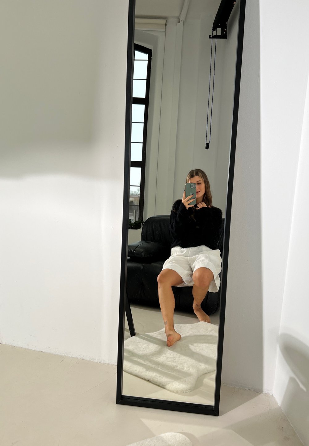 Specchio a figura intera con una cornice nera, che riflette una persona in un maglione nero e pantaloni bianchi, seduta su un divano in pelle nera.