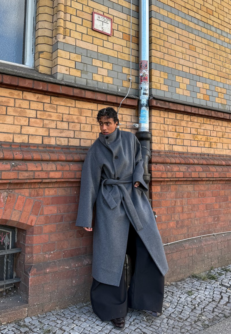 Personne portant un long manteau gris avec une ceinture, debout contre un mur en briques près d’une gouttière, regardant vers la gauche.