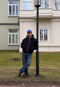 Un homme portant une casquette bleue LA, une veste noire et un jean est appuyé contre un lampadaire sur l'herbe devant un bâtiment clair avec des fenêtres.