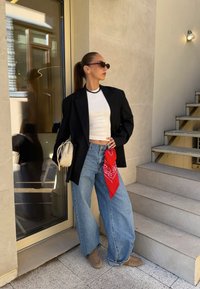 Fekete csíkos zakó, fehér cropped top, széles lábú kék farmer piros bandanával, bézs kézi táska, napszemüveg és bézs slip-on cipők. Kültéri környezet.