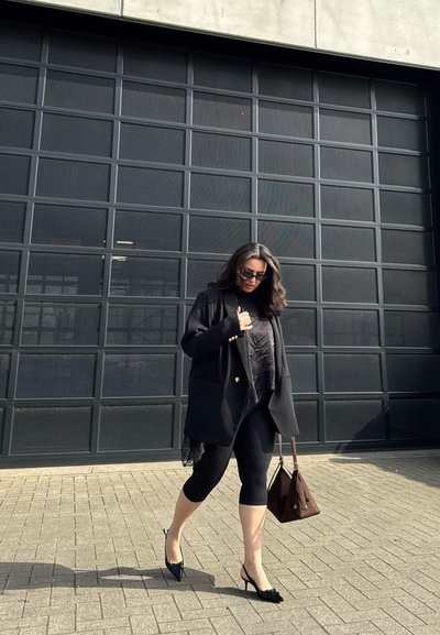 Mujer con blazer negro, leggings y tacones caminando al aire libre con bolso marrón, frente a una gran puerta de garaje oscura con patrón de rejilla.