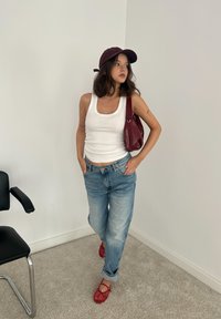 Top blanco sin mangas, jeans amplios de color azul claro, zapatos de malla rojos y una gorra color burdeos; sosteniendo un bolso de cuero burdeos. Fondo liso. Silla negra cercana.