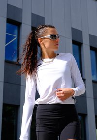 Naine põimitud hobusesabaga, seljas valge pikkade varrukatega spordisärk, mustad lühikesed püksid, päikeseprillid ja kuldne kett, seisab tänapäevase hoone ees.