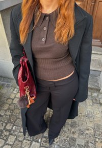 Brun stickad topp med krage, svarta vida byxor och en glänsande vinröd väska med en pälsboll-nyckelring. Bakgrund av stenlagd mark.