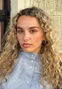 Młoda kobieta z długimi, kręconymi blond włosami, nosząca złote kolczyki koła i niebieską koszulę w paski na guziki, stojąca na tle teksturowanej ściany.