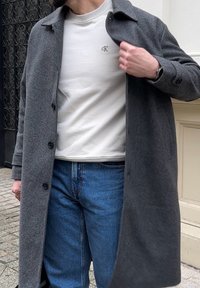 Cappotto grigio in misto lana con vestibilità su misura, dotato di chiusura con bottoni e colletto con patta, indossato sopra un maglione chiaro e jeans blu.
