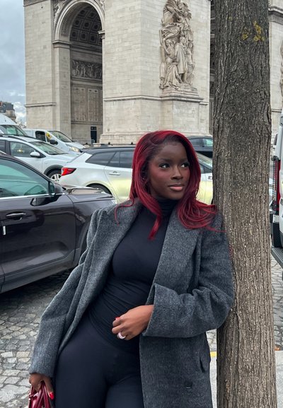 Mujer con cabello rojo que lleva un abrigo gris y un conjunto negro apoyada contra un árbol cerca del Arco de Triunfo con coches estacionados al fondo.