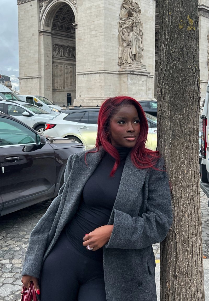 Mujer con cabello rojo que lleva un abrigo gris y un conjunto negro apoyada contra un árbol cerca del Arco de Triunfo con coches estacionados al fondo.