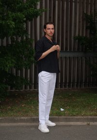 Čierne pruhované nadrozmerné tričko, biele nohavice a biele tenisky. Muž stojí oproti drevenému plotu s zeleňou na pozadí.