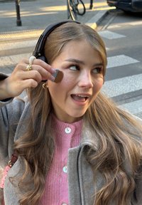 Jonge vrouw met lang haar die een grijze jas draagt, brengt met een kwast make-up aan op haar wang op een stoep in de stad.