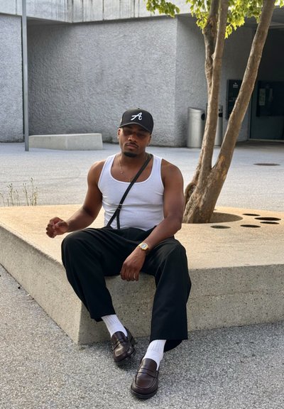 Gorra negra, camiseta blanca sin mangas, pantalones negros, mocasines marrones, calcetines blancos. Sentado en un banco de concreto con un árbol de fondo.