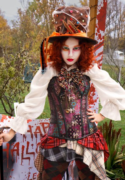 Persona con cabello rizado rojo y maquillaje facial blanco que lleva un sombrero de copa steampunk decorado, corsé colorido y falda con capas y estampados al aire libre.