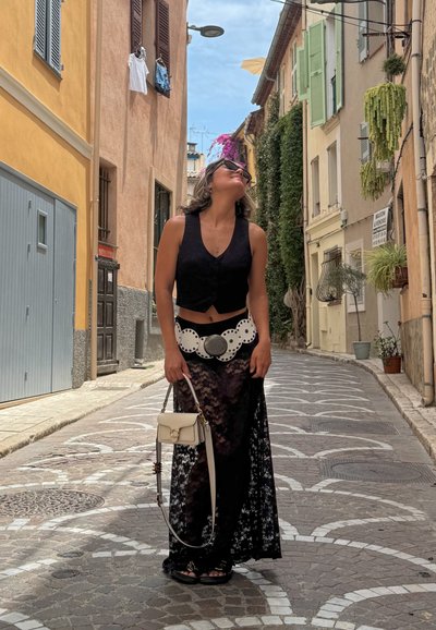 Top corto negro, falda maxi de encaje, cinturón blanco con recortes circulares y pequeño bolso crema; el fondo muestra una estrecha calle de adoquines.
