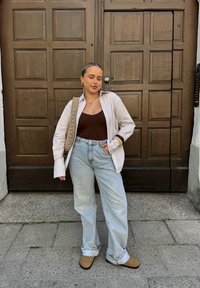 Gestreiftes Hemd, brauner Tanktop, hellblaue weite Jeans und beigefarbene Stiefeletten. Braune Tasche mit dekorativen Nieten. Holzverkleideter Hintergrund.