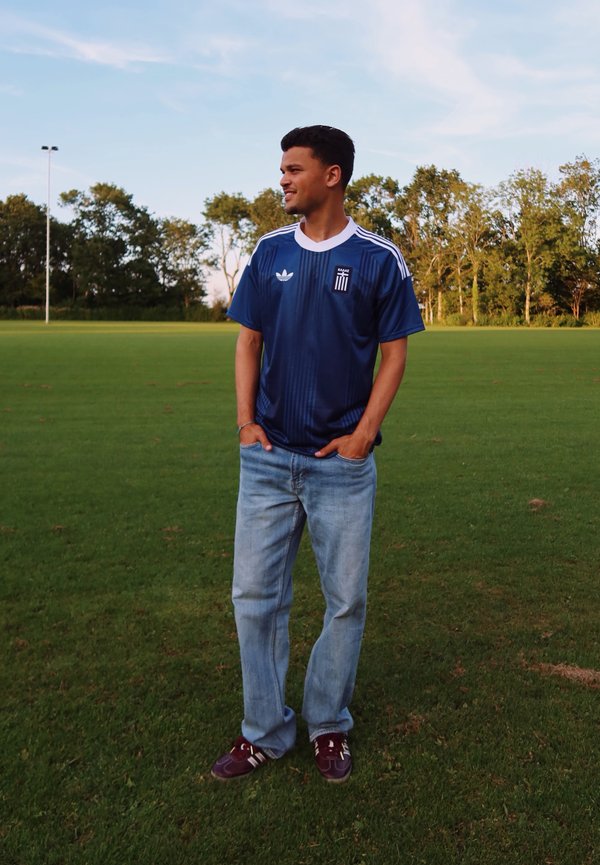 Jeune homme portant un maillot de sport bleu et un jean, debout dans un champ herbeux avec des arbres et un ciel bleu en arrière-plan.