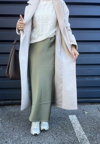 Κρέμα καφέ παλτό πάνω από ένα λευκό πουλόβερ με σχέδιο καρδιών, ελαιόχρωμη σατέν φούστα και λευκά αθλητικά. Ορατή τσάντα ώμου καφέ και χειρονομία χεριού.