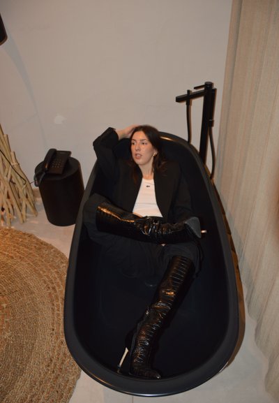 Mujer vestida con blazer negra, camiseta blanca y botas negras hasta la rodilla, reclinada en una bañera independiente negra en el interior.
