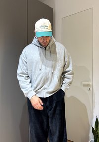 Homme portant un sweat à capuche gris clair zippé, un pantalon en velours côtelé noir et une casquette blanche et verte, debout à l'intérieur près d'une porte fermée et d'une plante.