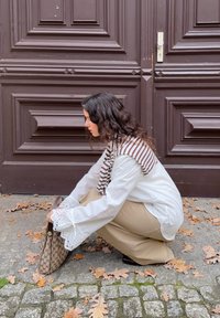 Hvit bluse med blonder i ermene, brunt og hvitt stripet skjerf, beige bukser og et mønstret håndveske, hukende på brostein ved siden av blader.