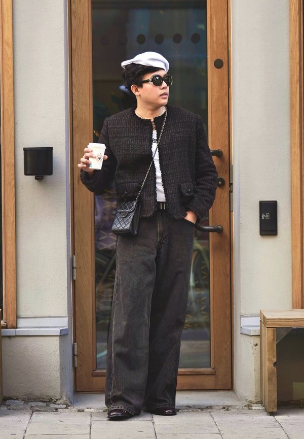 Persona que lleva una boina blanca, gafas de sol negras, una chaqueta negra y pantalones anchos negros, sosteniendo una taza de café frente a una puerta de madera.