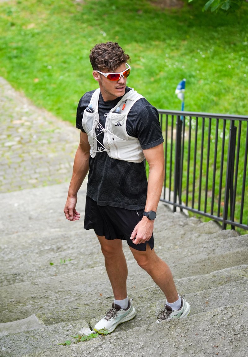 Homme portant une tenue de sport et des lunettes de soleil debout sur des marches en pierre extérieures, tenant la rampe, regardant vers sa gauche, avec de l'herbe en arrière-plan.